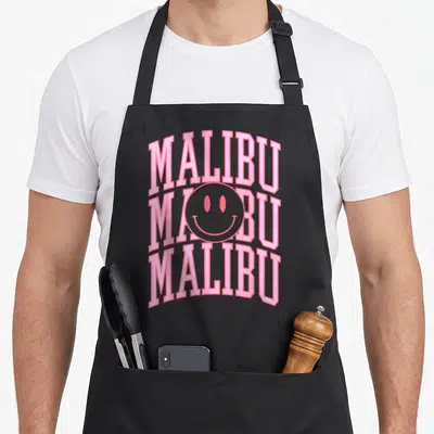 Funny preppy malibu apron - Pink MALIBU text stacked vertically with a centered smiley face overlay — shown on a black cotton canvas apron against a white background