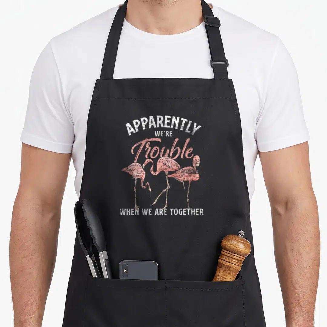 Hobby pink flamingo apron - Distressed four pink flamingos with apparently we're trouble when we are together text — shown on a black cotton canvas apron against a white background