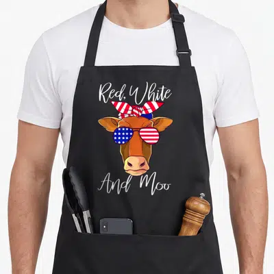 Patriotic cow apron - Brown cartoon cow head with flag sunglasses and striped bandana under Red White And Moo text — shown on a black cotton canvas apron against a white background