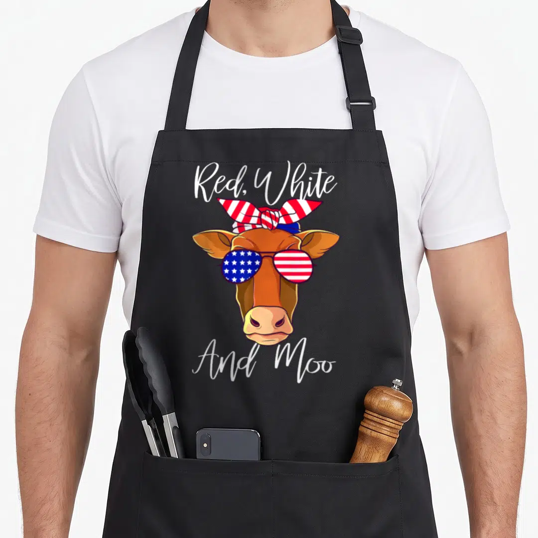 Patriotic cow apron - Brown cartoon cow head with flag sunglasses and striped bandana under Red White And Moo text — shown on a black cotton canvas apron against a white background