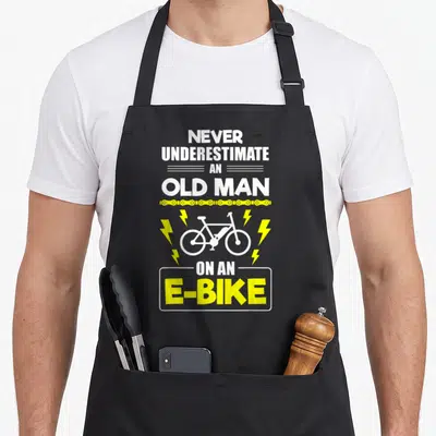 Ebike underestimate bike apron - White stacked text with yellow chain and e-bike silhouette flanked by lightning bolts — shown on a black cotton canvas apron against a white background