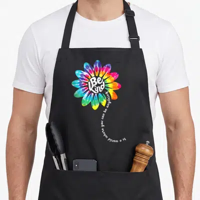 Flower tie-dye apron - Tie-dye flower with Be Kind text and curving sentence in white, blue, pink, yellow, green — shown on a black cotton canvas apron against a white background