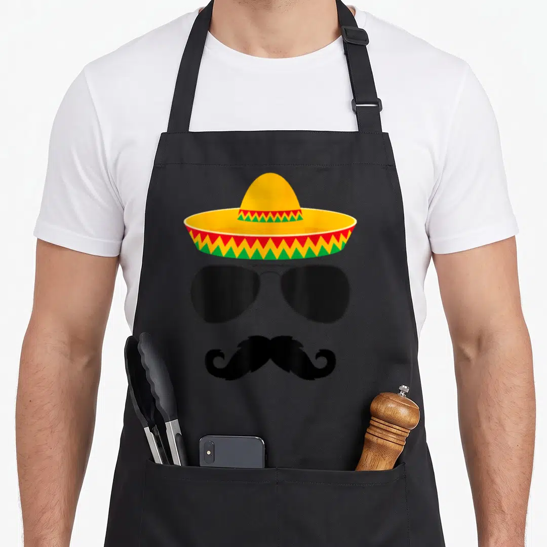 Cinco face apron - Yellow sombrero with red and green zigzags, black sunglasses, and curled mustache in cartoon style — shown on a black cotton canvas apron against a white background