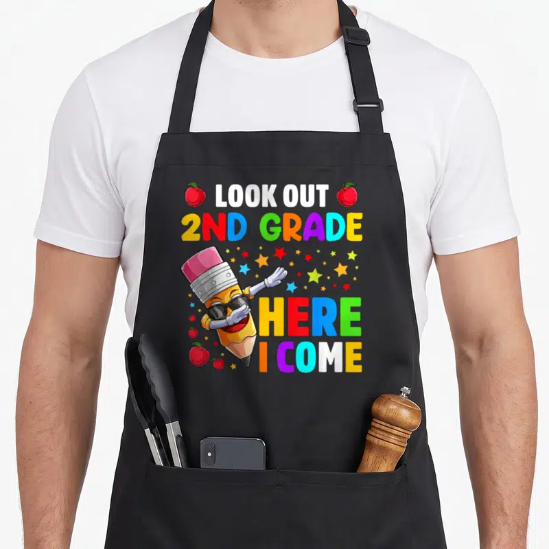 Come 2nd apron - Cartoon pencil with sunglasses and rainbow text reading LOOK OUT 2ND GRADE HERE I COME with apples — shown on a black cotton canvas apron against a white background