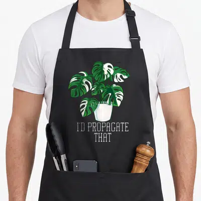 Propagate plant lady apron - Cartoon monstera in white pot with text I'D PROPAGATE THAT — shown on a black cotton canvas apron against a white background