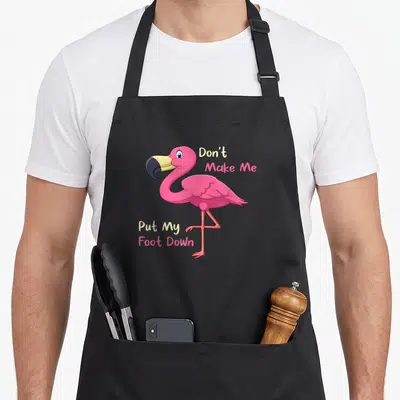 Foot tropical pink flamingo apron - Pink cartoon flamingo standing on one leg with text Don't Make Me Put My Foot Down — shown on a black cotton canvas apron against a white background