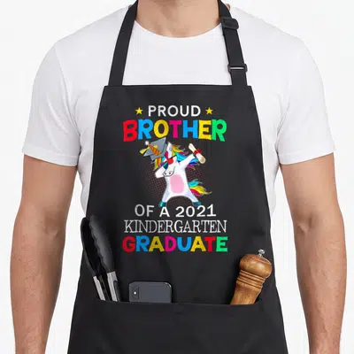 Brother unicorn aprons - Dabbing unicorn with cap diploma and proud brother kindergarten graduate text — shown on a black cotton canvas apron against a white background