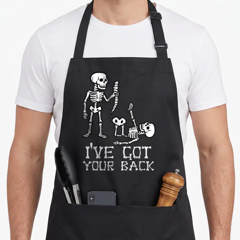 Halloween costume skeleton apron - Two cartoon skeletons with bone text reading I've Got Your Back — shown on a black cotton canvas apron against a white background