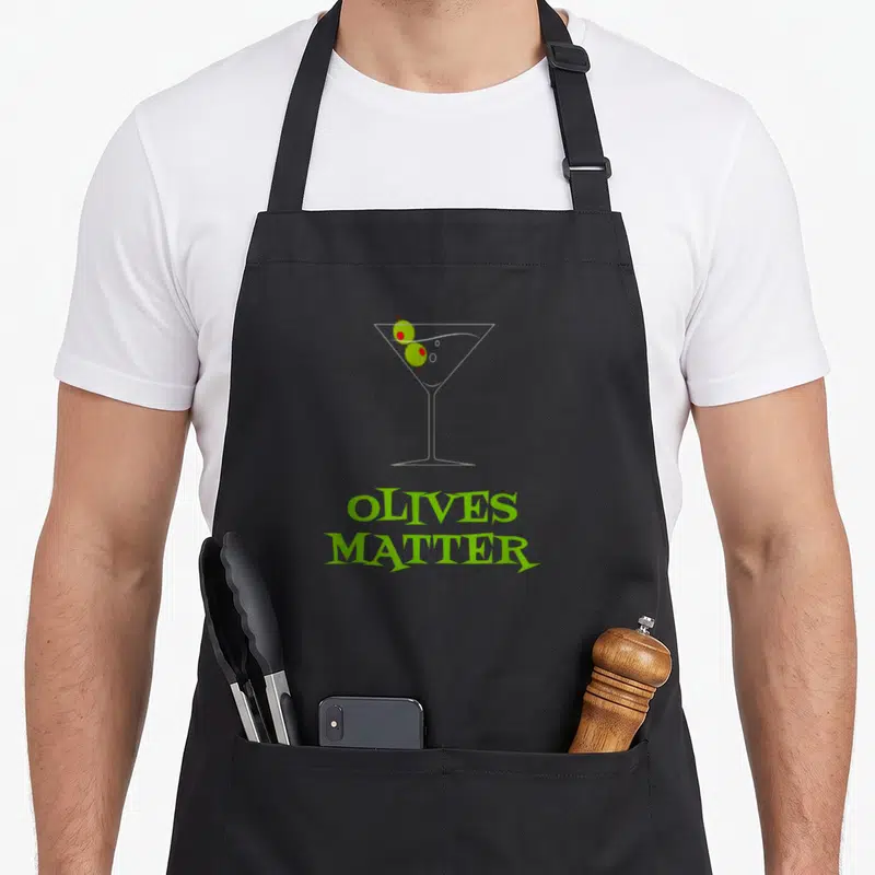 Martini cosmopolitan apron - Minimalist martini glass with two green olives and red picks above stacked green text — shown on a black cotton canvas apron against a white background