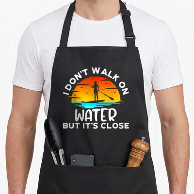 Paddleboarding paddleboard girl apron - Retro sunset paddleboarder silhouette with text I don't walk on water but it's — shown on a black cotton canvas apron against a white background