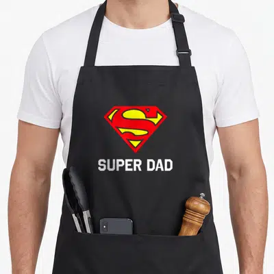 Funny family father day apron - Red yellow shield logo above white SUPER DAD text — shown on a black cotton canvas apron against a white background