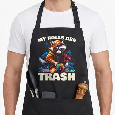 Tabletop game gaming apron - Raccoon in orange hoodie holding d20 die with MY ROLLS ARE TRASH text — shown on a black cotton canvas apron against a white background