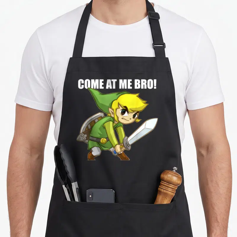 Come legend apron - Link holding sword and shield with white COME AT ME BRO! text above — shown on a black cotton canvas apron against a white background