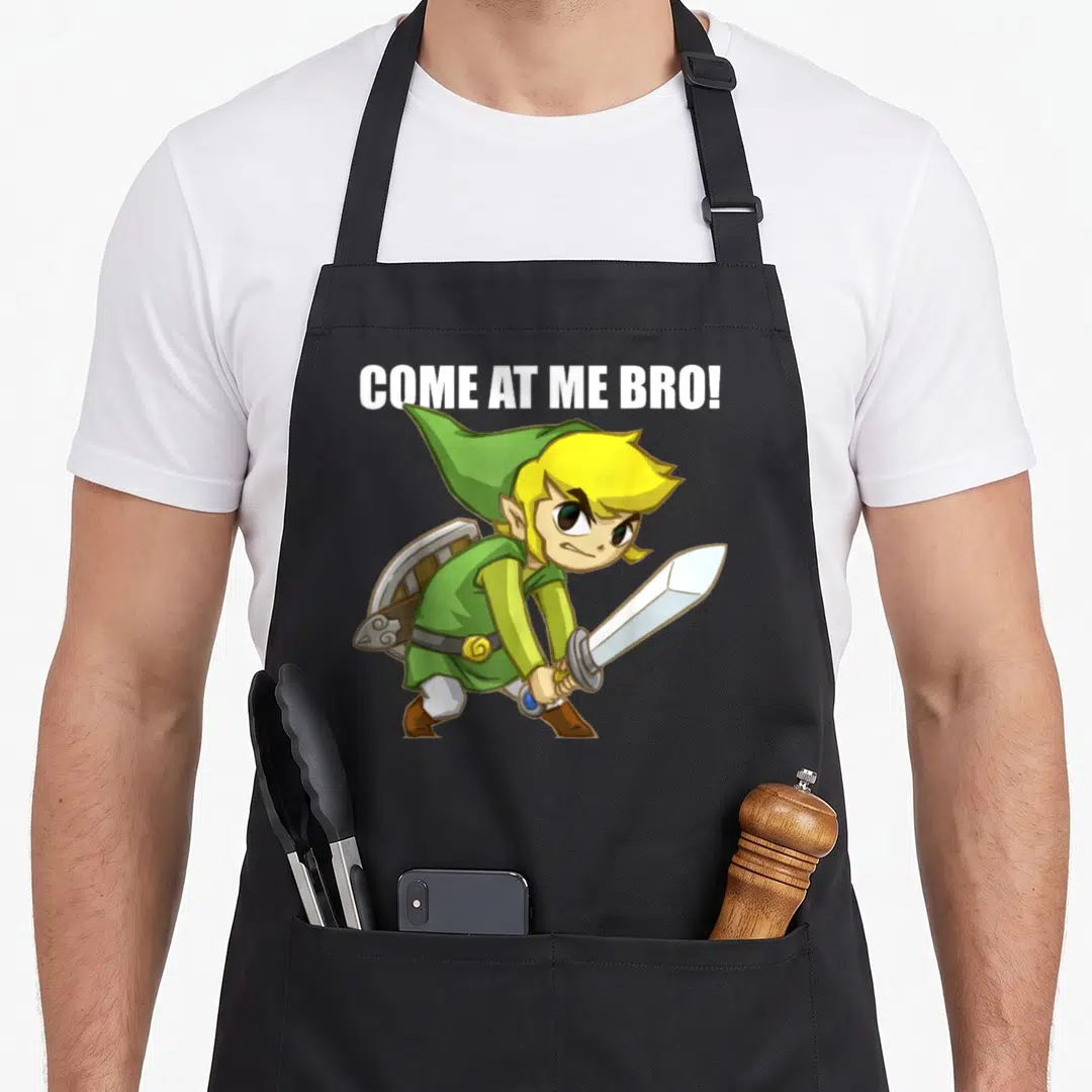 Come legend apron - Link holding sword and shield with white COME AT ME BRO! text above — shown on a black cotton canvas apron against a white background