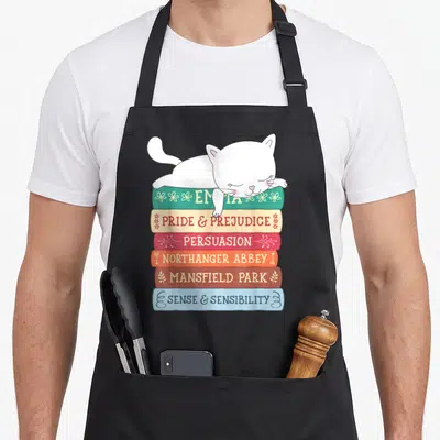 Book jane austen apron - Sleeping white cat on stack of six books with spines labeled Emma and Pride & Prejudice — shown on a black cotton canvas apron against a white background