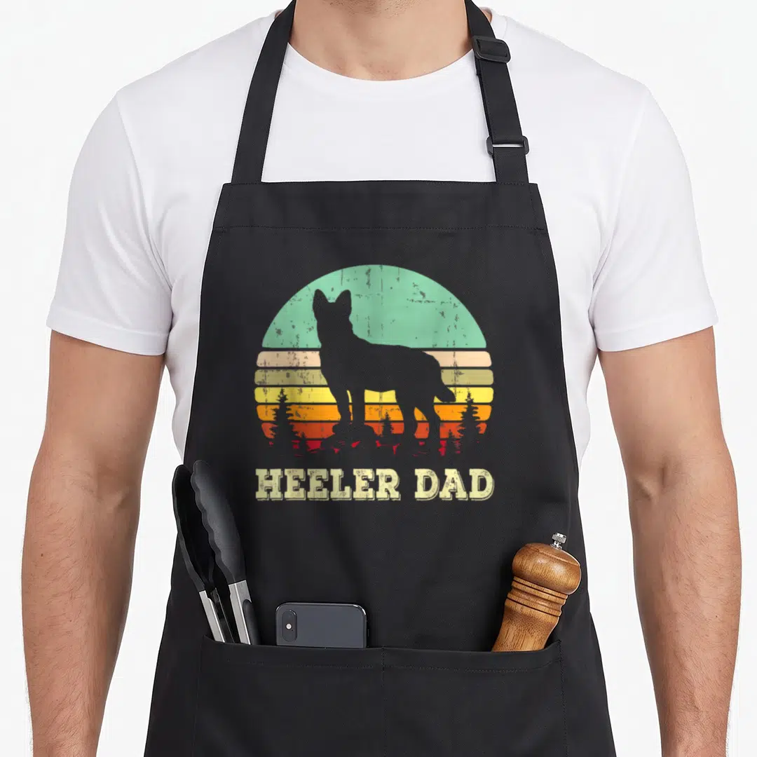Cattle dog kitchen apron - Grey dog silhouette against retro striped sun with HEELER DAD text below — shown on a black cotton canvas apron against a white background