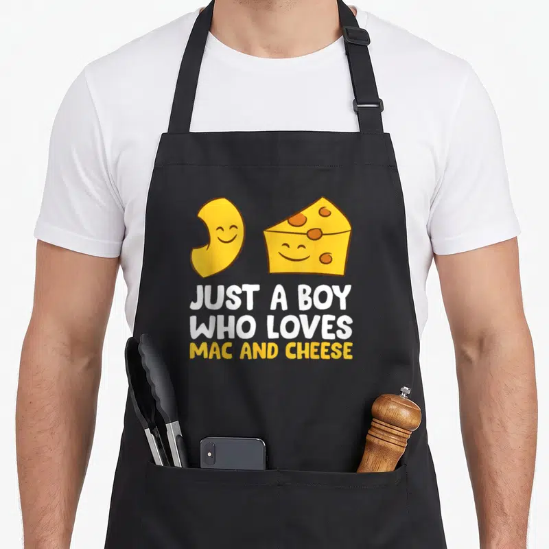 Funny macaroni mac apron - Cartoon cheese and smiling noodle above JUST A BOY WHO LOVES MAC AND CHEESE text — shown on a black cotton canvas apron against a white background