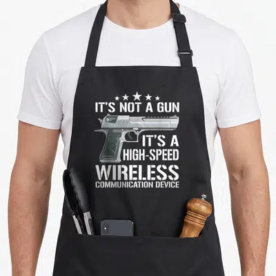 Gun meme apron - Realistic silver pistol with white stencil text reading IT'S NOT A GUN HIGH-SPEED WIRELESS — shown on a black cotton canvas apron against a white background