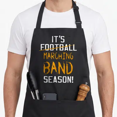 Marching band football apron - White and orange stacked text reading It's Marching Band Season with Football crossed out — shown on a black cotton canvas apron against a white background