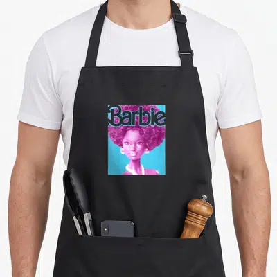 Funny afro doll apron - Pink doll with large curly hair and grey Barbie text on blue background — shown on a black cotton canvas apron against a white background