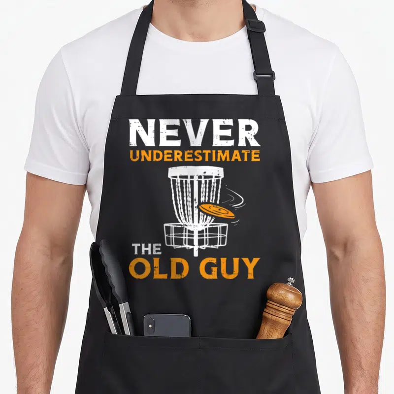 Boredkoalas golf apron - Distressed text Never Underestimate Old Guy above and below white disc golf basket with flying — shown on a black cotton canvas apron against a white background