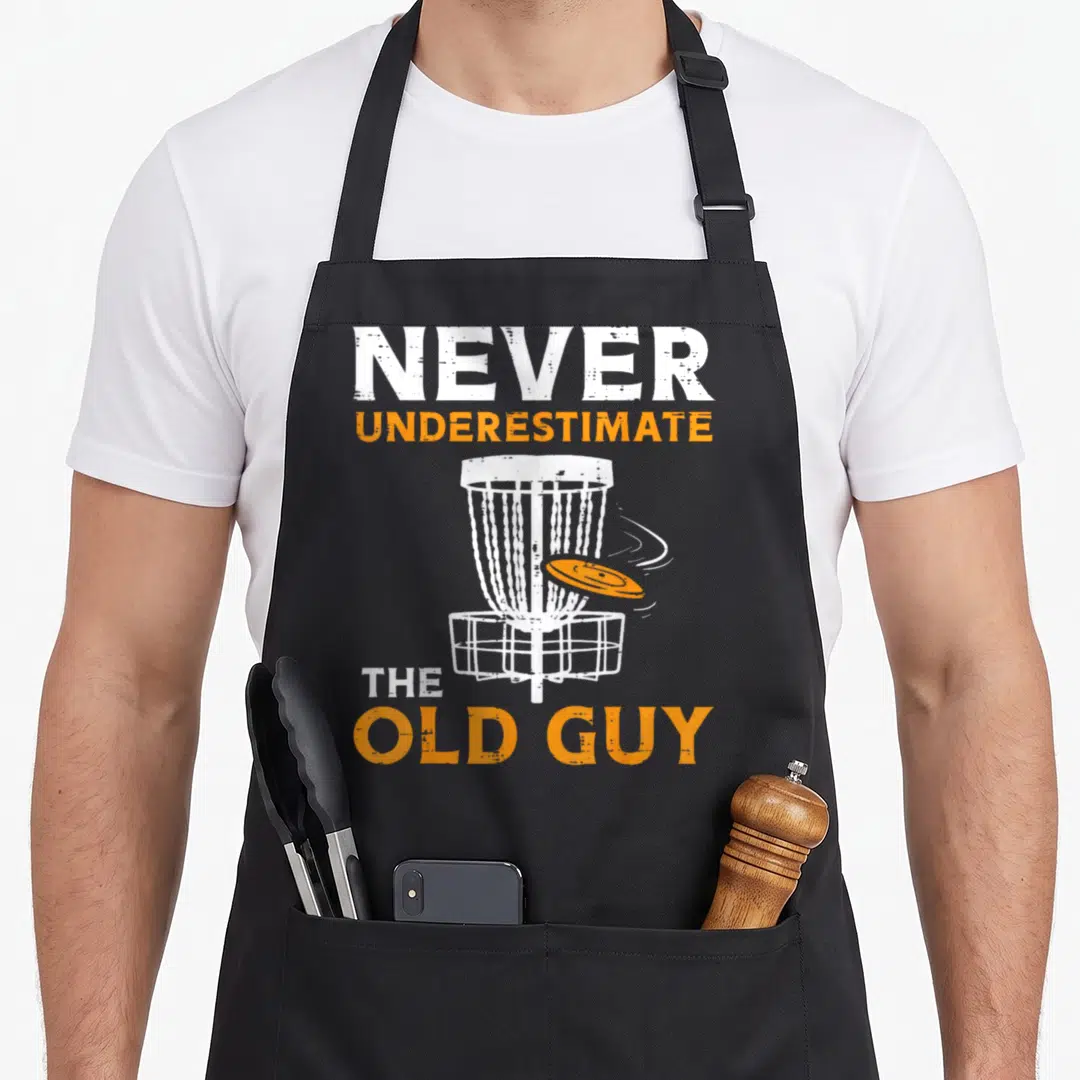 Boredkoalas golf apron - Distressed text Never Underestimate Old Guy above and below white disc golf basket with flying — shown on a black cotton canvas apron against a white background