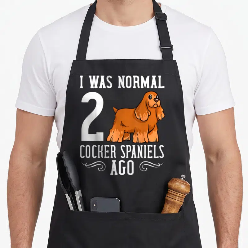 Puppy cocker spaniel apron - Orange cartoon cocker spaniel beside white number 2 and text I was normal ago — shown on a black cotton canvas apron against a white background