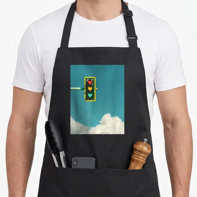 Indie 90s apron - Yellow traffic light with red, yellow, and green heart signals against teal sky and white clouds — shown on a black cotton canvas apron against a white background