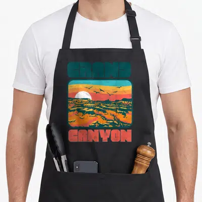 Grand 80s apron - Retro Grand Canyon landscape with sun birds and text in teal coral orange yellow — shown on a black cotton canvas apron against a white background