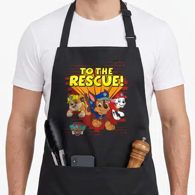 Rescue birthday crew apron - Three cartoon puppies running under TO THE RESCUE! text with brick wall background — shown on a black cotton canvas apron against a white background