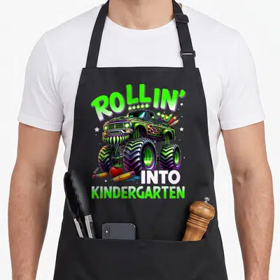Rollin truck apron - Green monster truck with ROLLIN INTO KINDERGARTEN text and scattered pencils — shown on a black cotton canvas apron against a white background