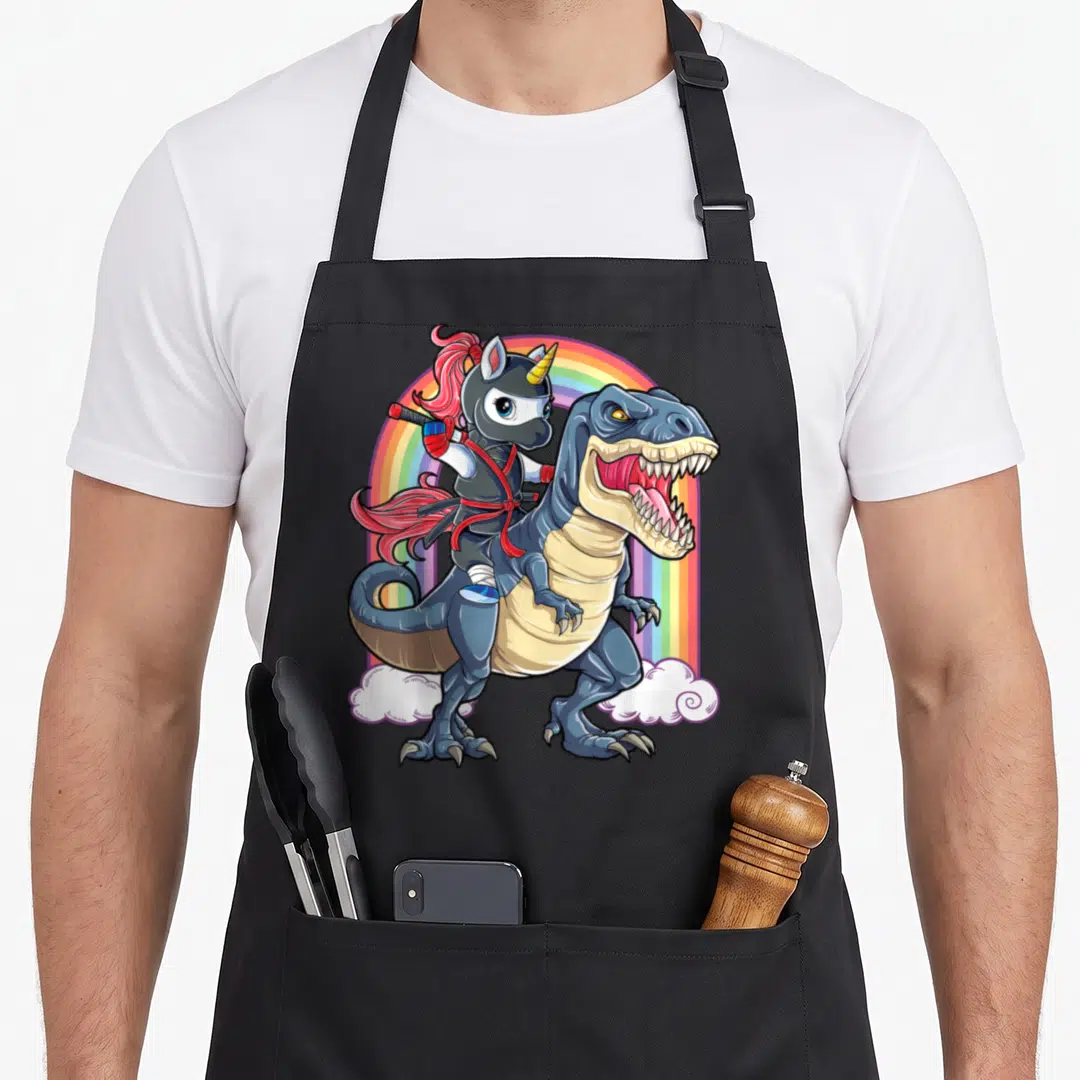 Ninja dinosaur apron - Cartoon unicorn ninja riding blue T-Rex with rainbow background and clouds — shown on a black cotton canvas apron against a white background