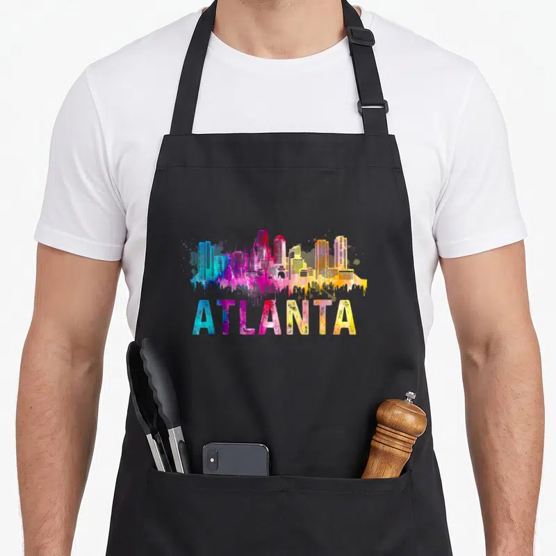 Georgia atlanta apron - Watercolor Atlanta skyline in blue purple pink yellow with paint drips over text — shown on a black cotton canvas apron against a white background