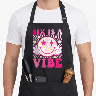 Smile birthday apron - Pink smiley face with daisies peace hands and text SIX IS A VIBE on checkerboard — shown on a black cotton canvas apron against a white background