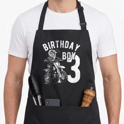 Motorcycle birthday apron - White distressed graphic of motocross rider and text BIRTHDAY BOY 3 — shown on a black cotton canvas apron against a white background