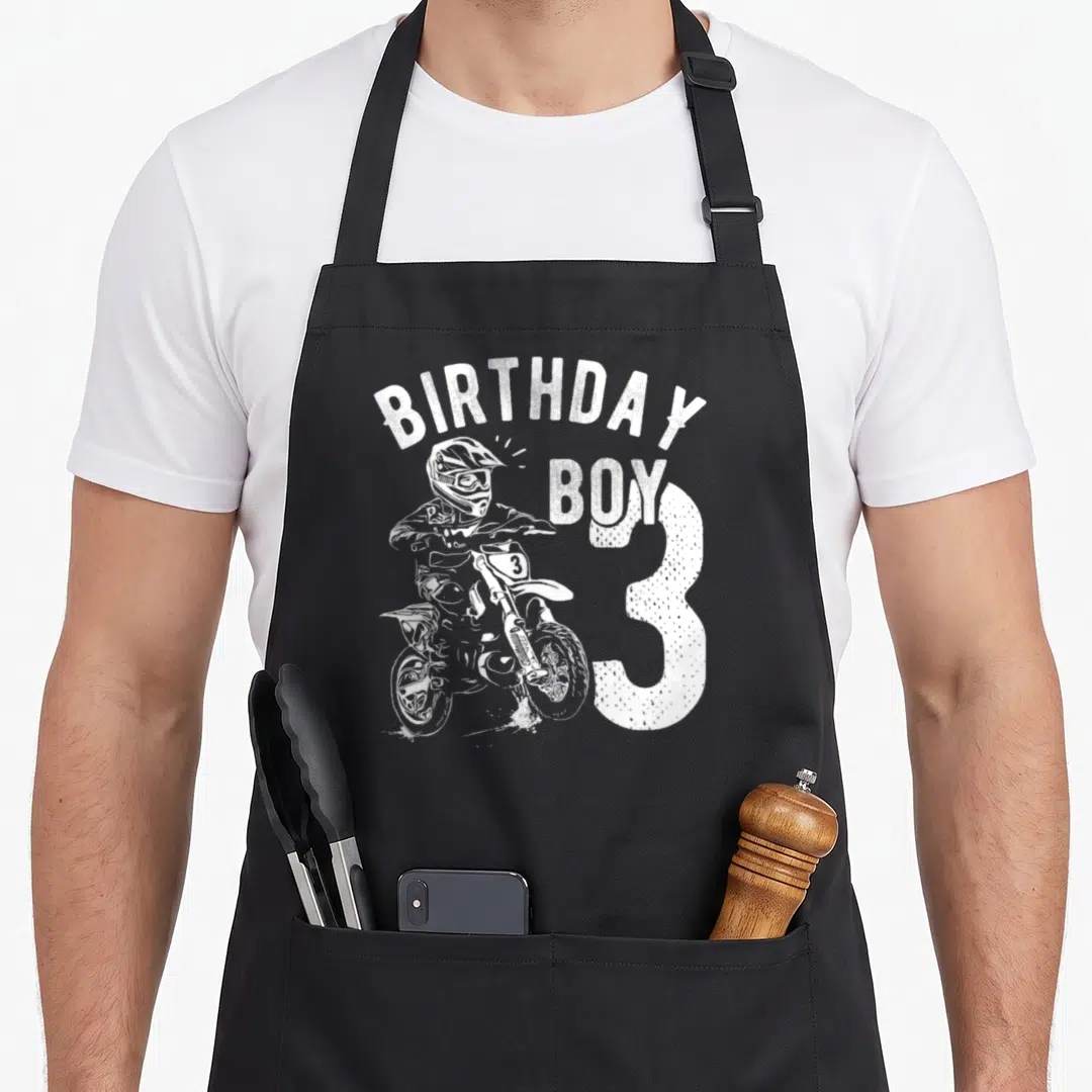 Motorcycle birthday apron - White distressed graphic of motocross rider and text BIRTHDAY BOY 3 — shown on a black cotton canvas apron against a white background