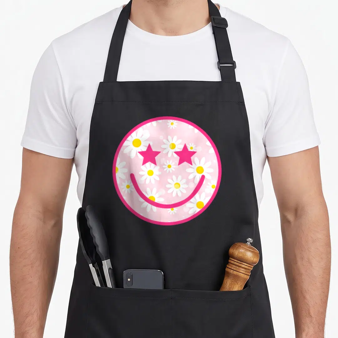 Daisy face on apron - Pink circle with star-eyed smiley face over white and yellow daisy floral pattern — shown on a black cotton canvas apron against a white background