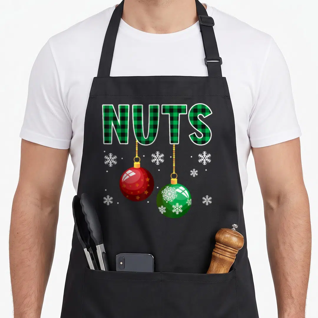 Chestnut couples christmas aprons - NUTS text in green plaid with hanging red and green ornaments and white snowflakes — shown on a black cotton canvas apron against a white background