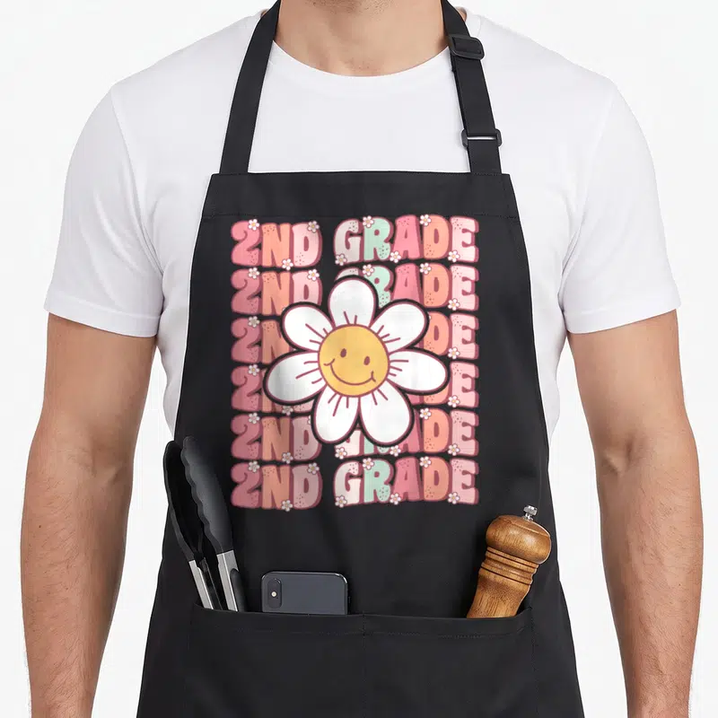 Groovy grade 2nd apron - Smiling daisy with white petals over six rows of 2ND GRADE text in pink, orange, white, yellow — shown on a black cotton canvas apron against a white background