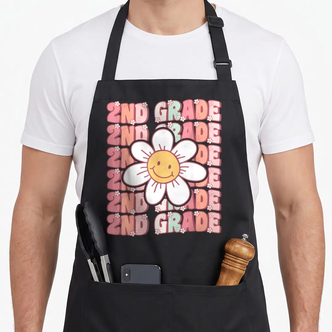 Groovy grade 2nd apron - Smiling daisy with white petals over six rows of 2ND GRADE text in pink, orange, white, yellow — shown on a black cotton canvas apron against a white background