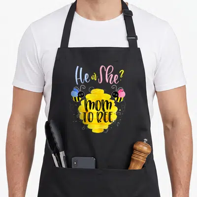 Reveal bee apron - He or She? text above yellow beehive with mom TO BEE and two balloon bees in pink and blue — shown on a black cotton canvas apron against a white background