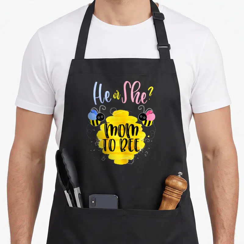 Reveal bee apron - He or She? text above yellow beehive with mom TO BEE and two balloon bees in pink and blue — shown on a black cotton canvas apron against a white background