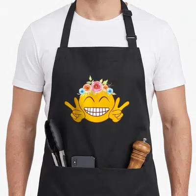 Emoticon face on apron - Yellow smiley face with watercolor flower crown and two peace sign hands — shown on a black cotton canvas apron against a white background