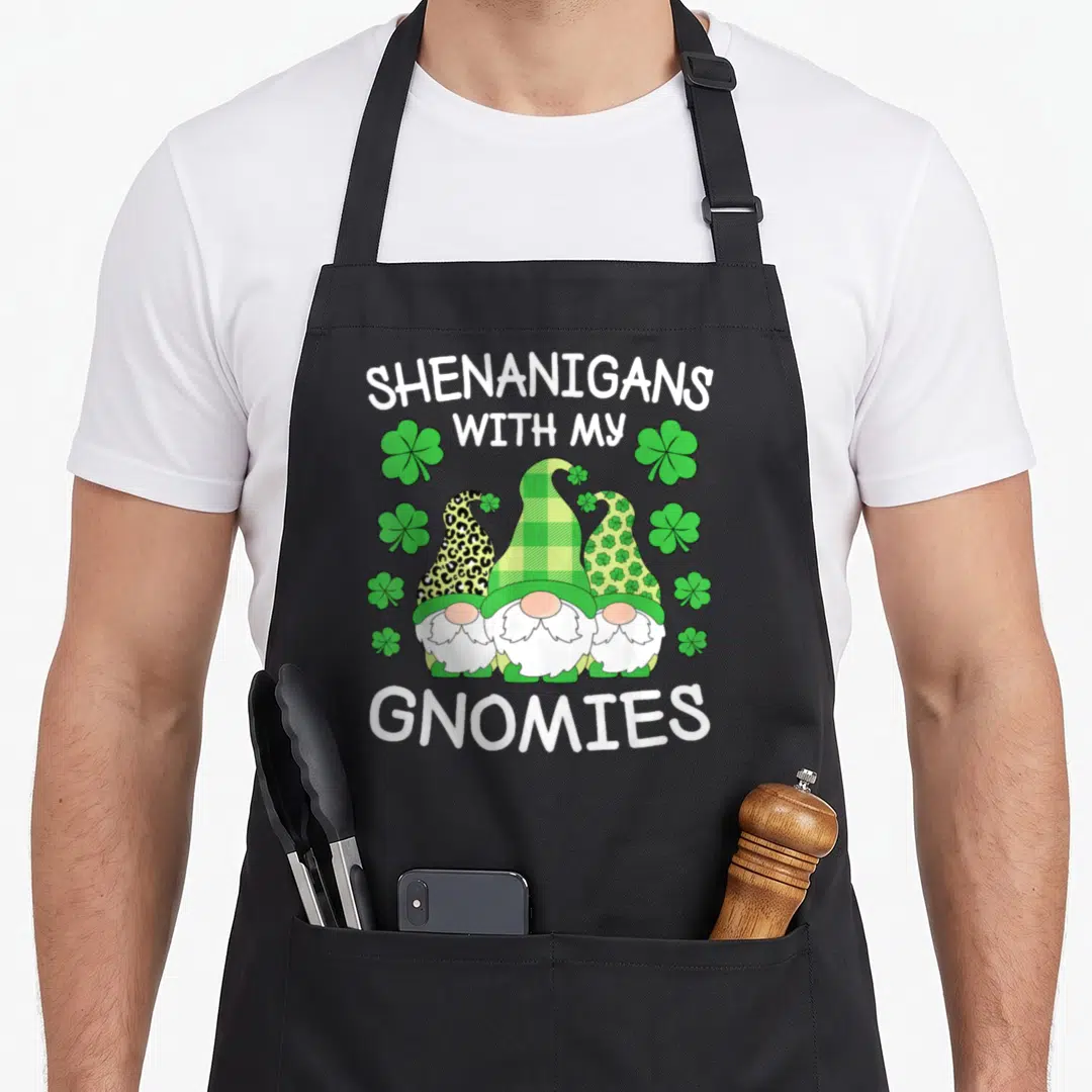 Gnomy st patrick day aprons - Three gnomes with patterned hats under arched text surrounded by green shamrocks — shown on a black cotton canvas apron against a white background