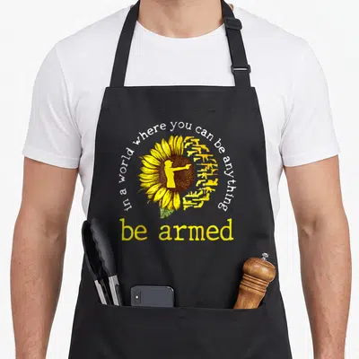 Armed sunflower apron - Sunflower with gun petals and silhouette holding a firearm under arched text — shown on a black cotton canvas apron against a white background