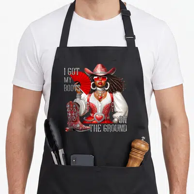 Hat western apron - Woman in red cowboy hat holding fan next to boot with text I GOT MY BOOTS ON THE GROUND — shown on a black cotton canvas apron against a white background
