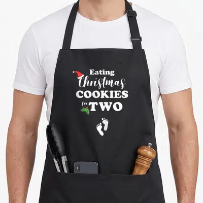 Pregnancy women's christmas aprons - Stacked text Eating Christmas Cookies for Two with Santa hat, holly, and baby — shown on a black cotton canvas apron against a white background