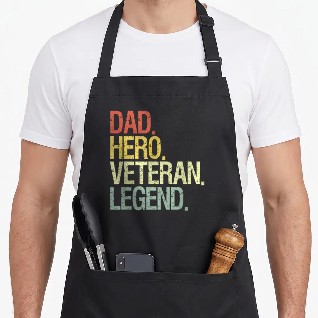 Cute veterans apron - Stacked DAD HERO VETERAN LEGEND text in red yellow and green distressed font — shown on a black cotton canvas apron against a white background