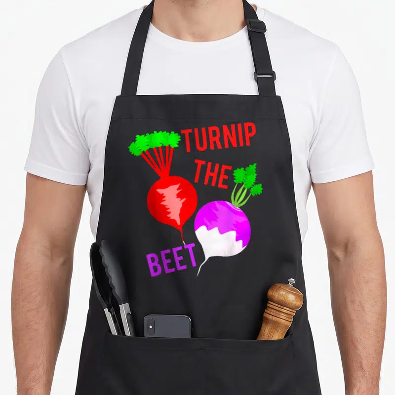 Turnip veggie apron - Cartoon red turnip and purple beet with text TURNIP THE BEET — shown on a black cotton canvas apron against a white background