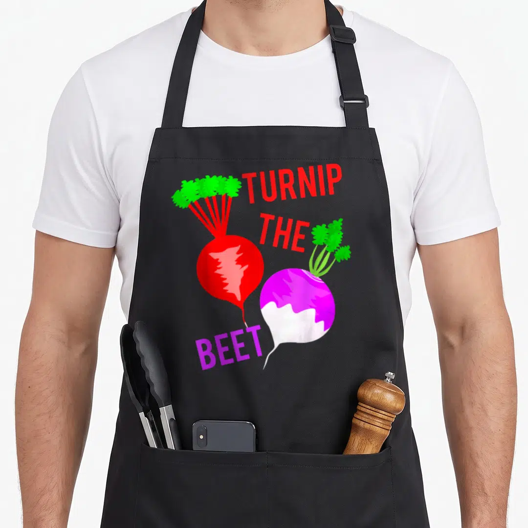 Turnip veggie apron - Cartoon red turnip and purple beet with text TURNIP THE BEET — shown on a black cotton canvas apron against a white background